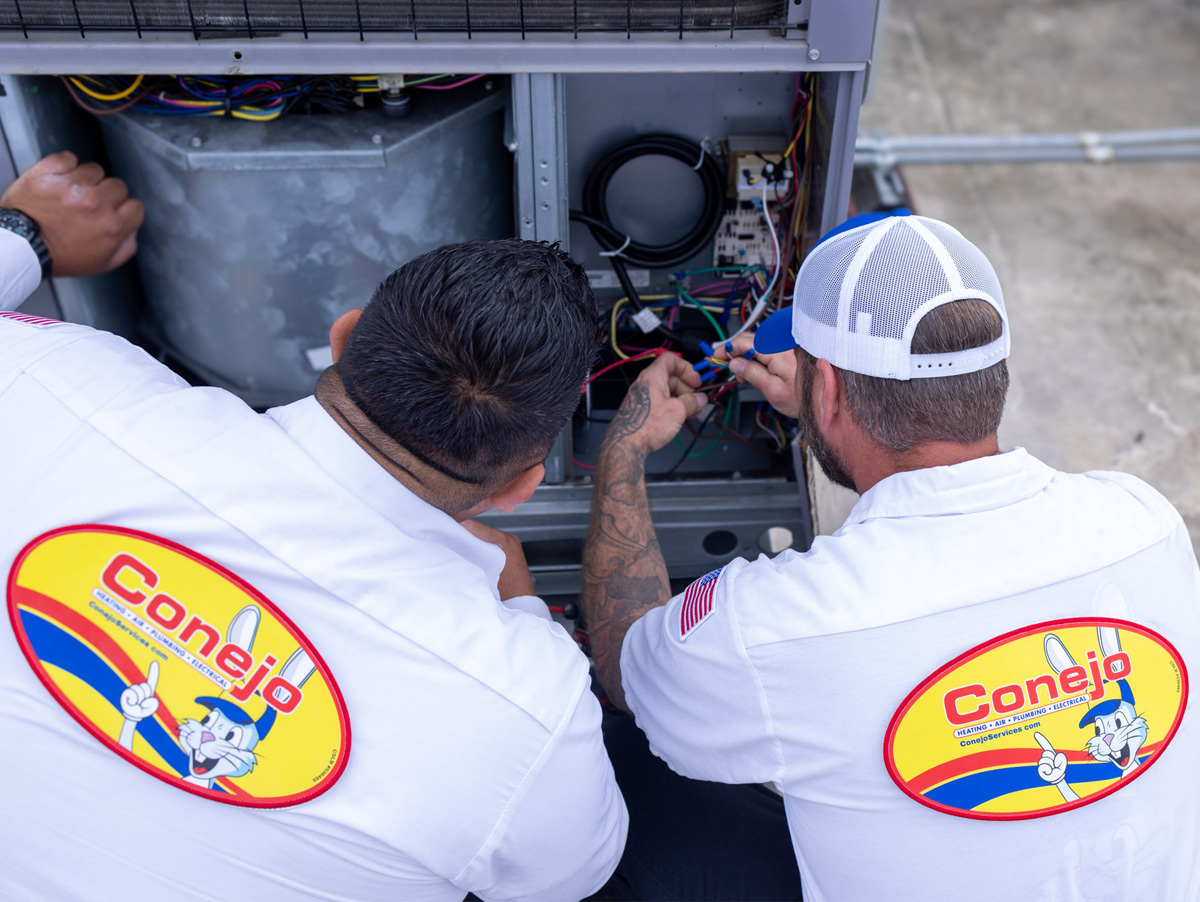 Two HVAC technicians repair an air conditioning unit, wearing Conejo Services shirts.