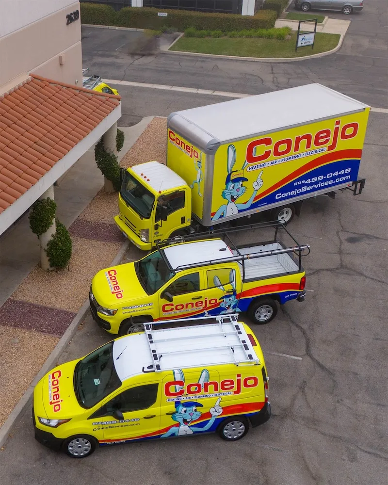 Three Conejo Services vehicles—a van, truck, and box truck—parked near a building. The vehicles are yellow with the Conejo logo.